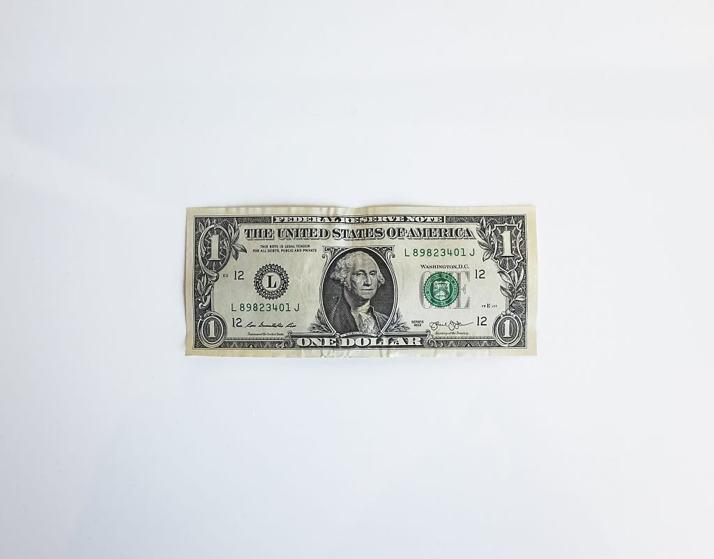 A photo of a US 1-Dollar bill sitting face-up on a flat, white surface.