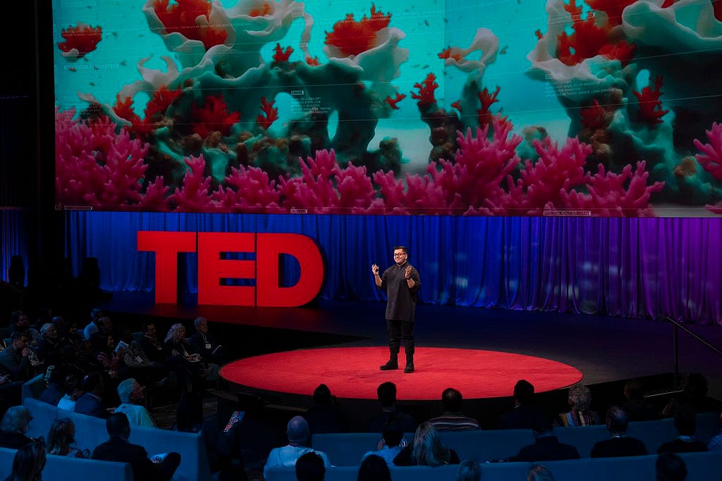 Refik Anadol stands on the TED stage, with 3D modeled corals of pink and blue surrounded by an ocean environment on the screen behind