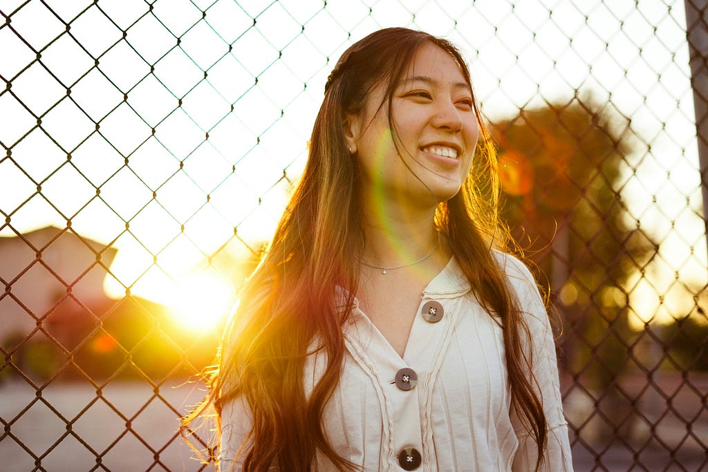 Smiling woman standing in front of chain-link fence, haloed by sunrise.
