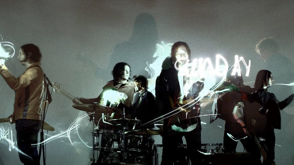 Shot from “Sunday Driver” music video; The Raconteurs band members playing their instruments and Jack White is shown in two places with the band and off to the left looking at white text that is written in the air around the band