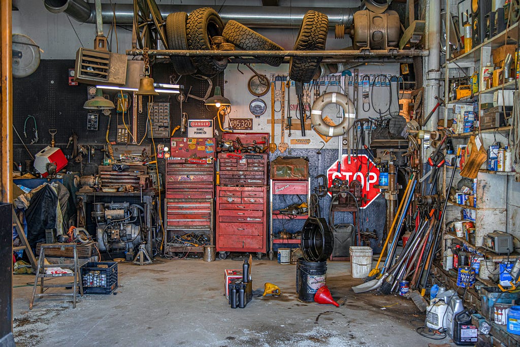 Cluttered garage space with some red toolboxes, pile of shovels, some old tires and a bunch of random bottles/cans and other clutter.