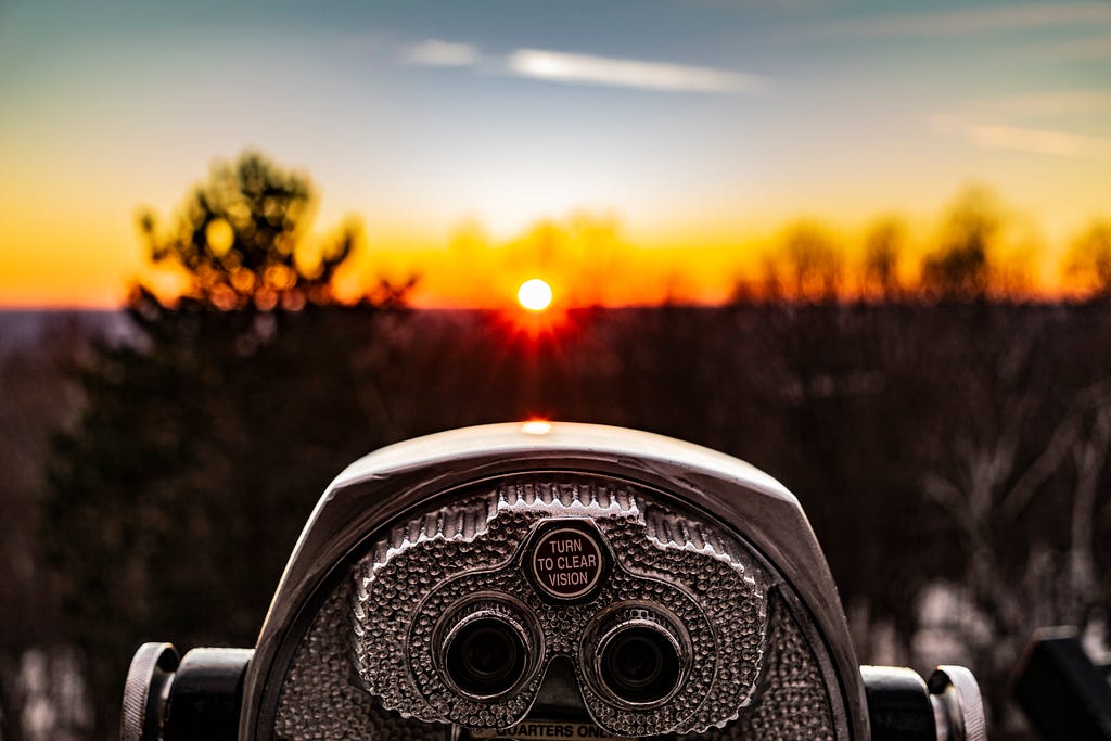 In-focus coin operated binoculars overlooking a blurry backdrop of the sunset and large trees.