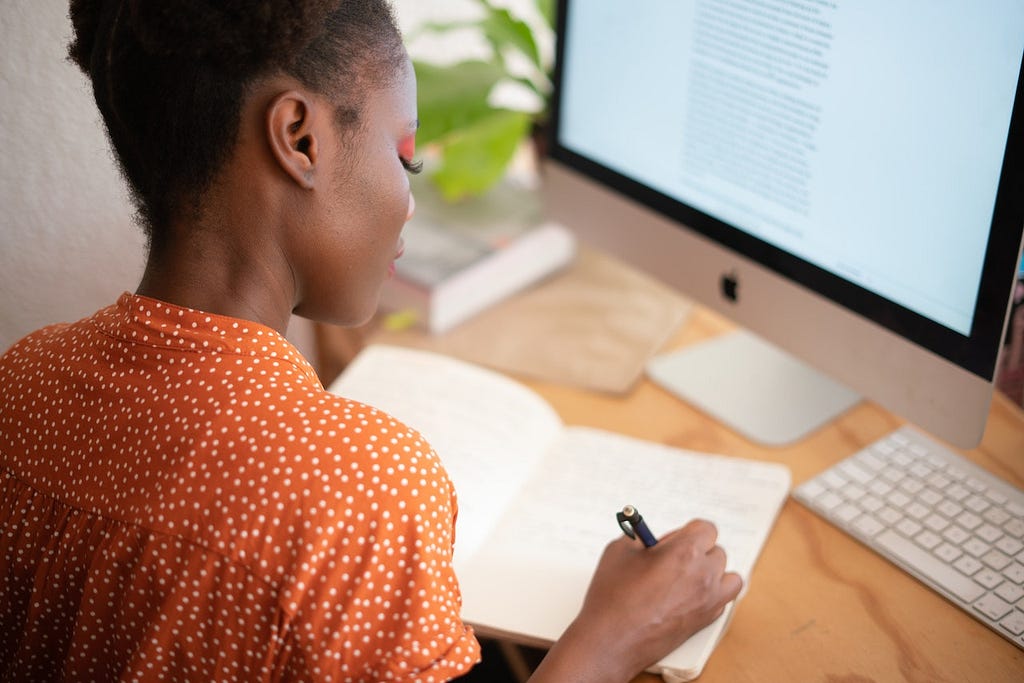 a woman looks down at her book as she writes in it in front of her computer, drafting a self-pleasure practice for her clients