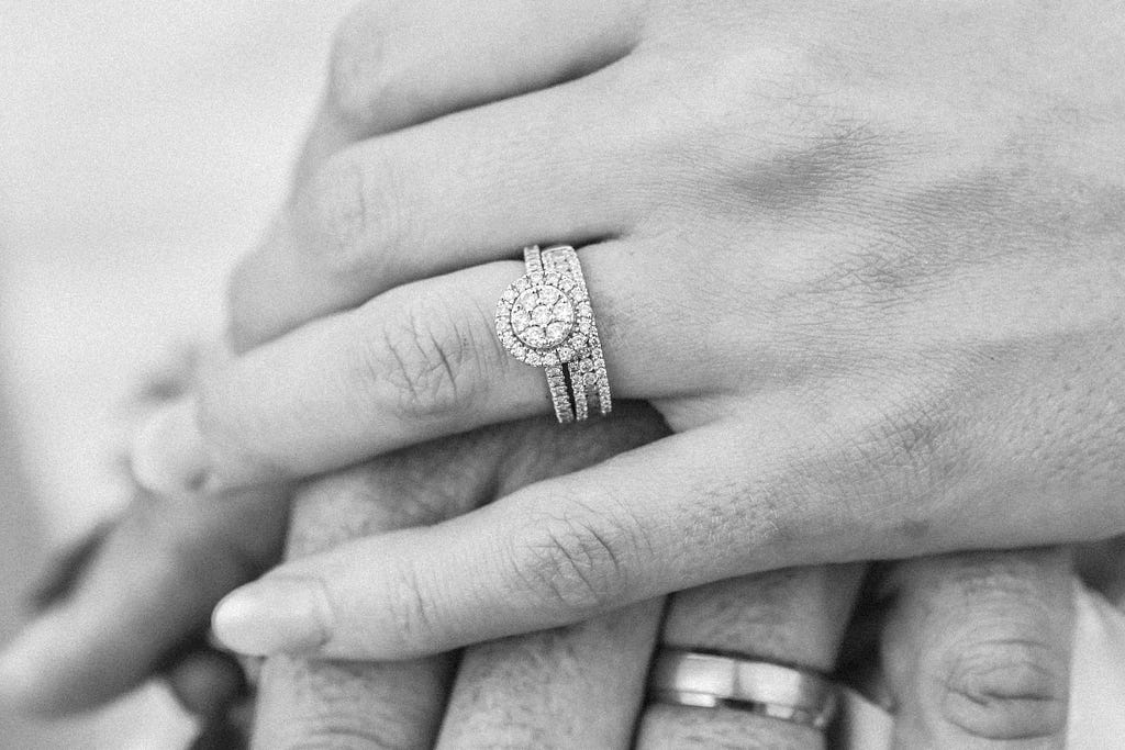 A woman’s hand atop man’s, displaying their engagement rings and wedding bands, symbolizing love and commitment.
