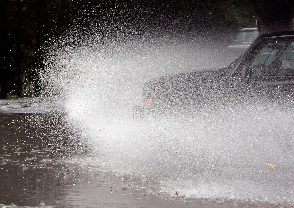 The front end of a dark car driving through a puddle and sending up a spray of water