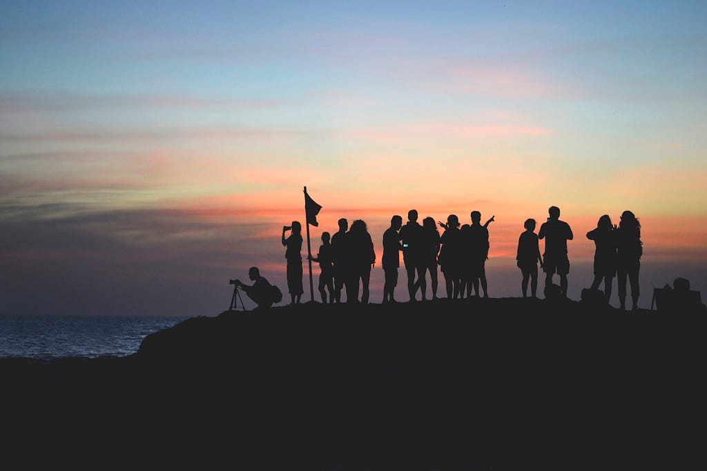 The image shows a group of people watching the sunset atop a rocky outcrop by the sea.