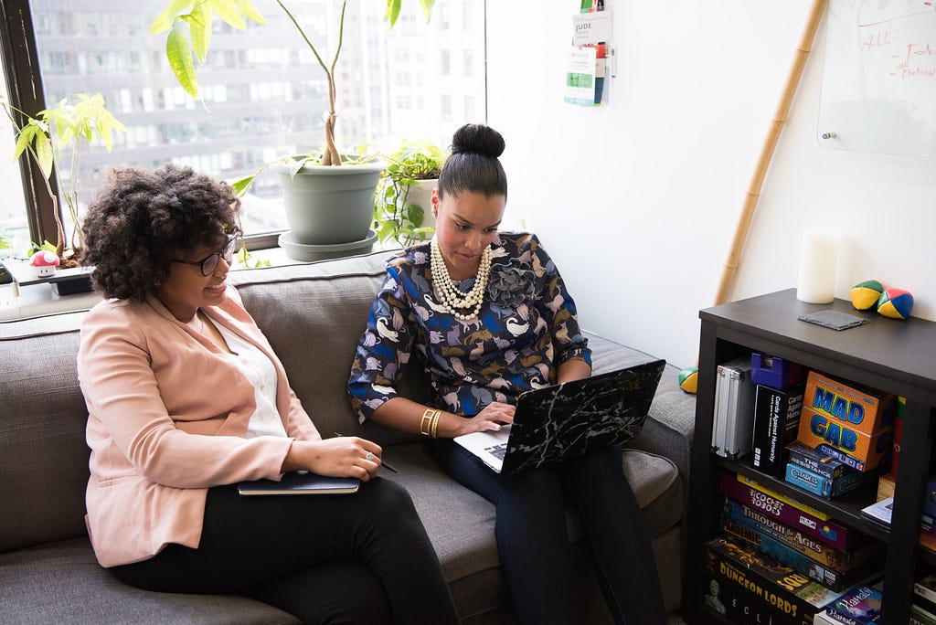 Two femme presenting people sitting on a sofa with a laptop on one of their laps looking over something together.