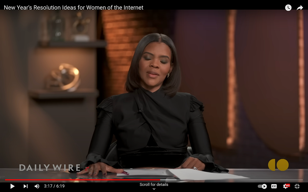 A screenshot from Candace Owens’s show on The Daily Wire. She wears a black dress that looks to be made of silk crepe. It has long sleeves with ruffle cuffs. There is also a bit of ruffle at the top of each shoulder. Fabric criss-crosses her chest and goes up to her neck in a kind of twist.