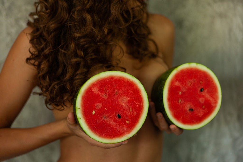 A woman holds two halves of a watermelon against her, like breasts. Cut open, as we are once we have breast cancer.