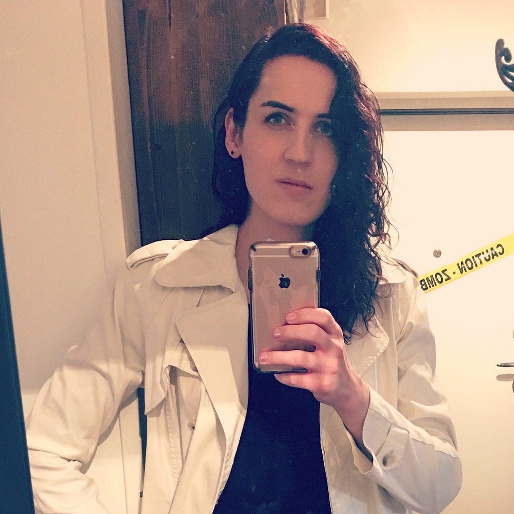 Mirror selfie of me in my late 20s. I have soft pink manicured nails, a rose gold iPhone, and I’m wearing a white trench coat.