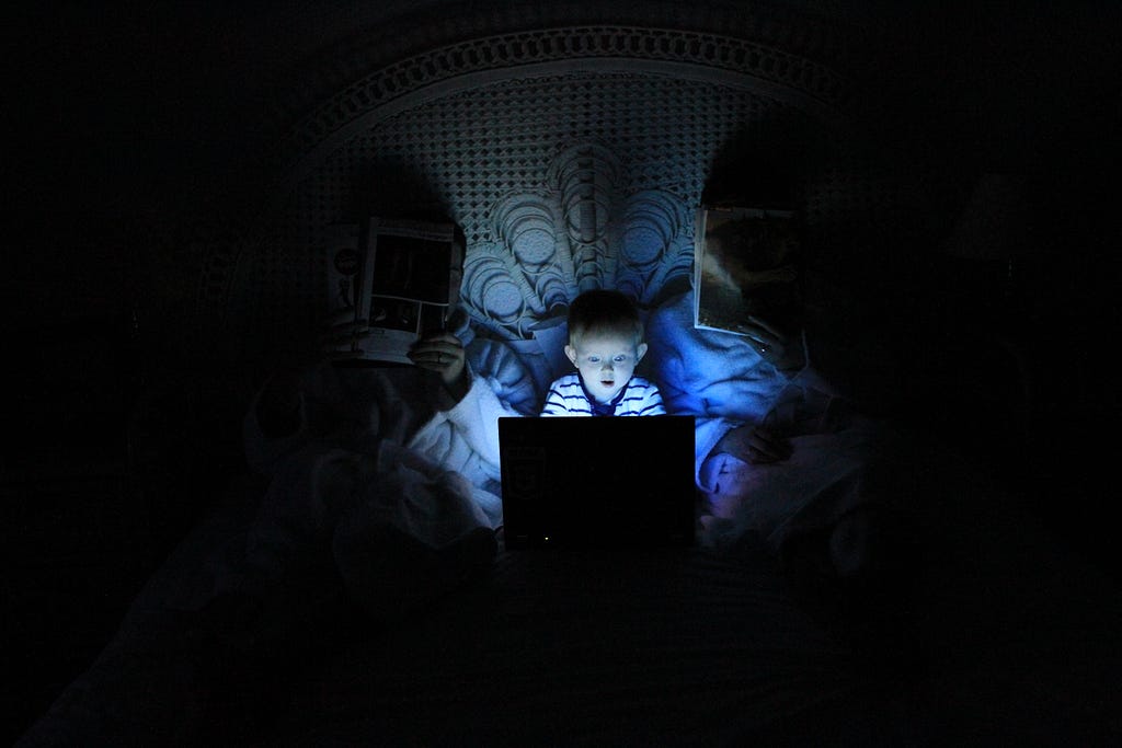Family sitting in bed in dark room. Parents reading books and child is staring in amazement at a computer screen.