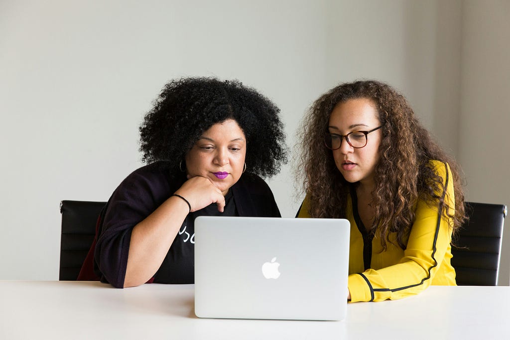 Two black women look at a laptop together in an office.