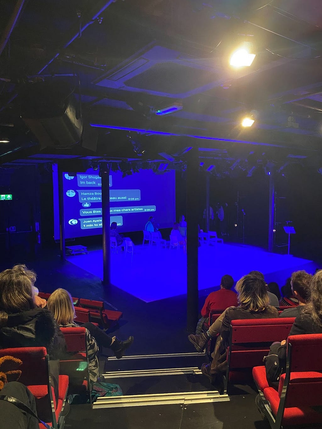 A blue-lit stage with a few people sat on chairs and a Whatsapp chat on the projection screen. Audience members on both sides of the image, sat on red seats.