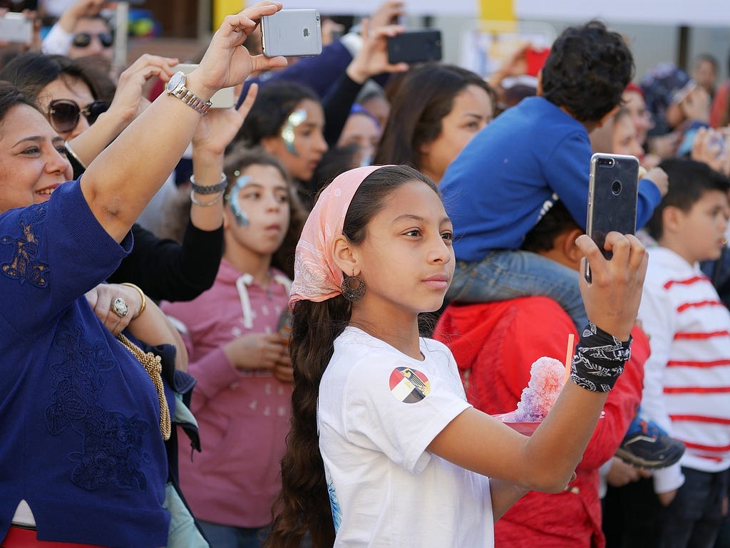 Registro de un público en el que varios individuos apuntan sus smartphones para registrar algo. En primer plano, una chica con un pañuelo sobre la cabeza y también empuñando su teléfono móvil.