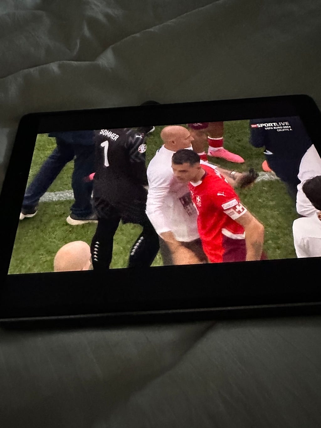 A tablet displays a scene of people on a sports field, with some wearing sports jerseys and others in white clothing, suggesting a post-game interaction.