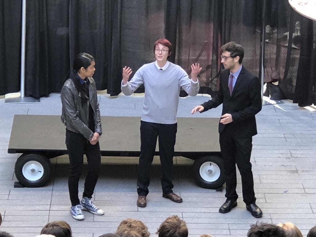 Three student actors portraying Bassiano, Antonio, and Shylock stand on a makeshift stage. Antonio holds up his hands and speaks while Bassiano and Shylock listen.