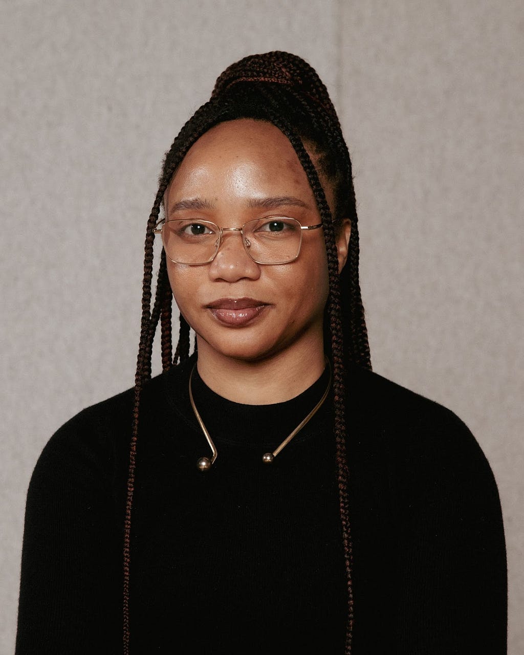 A portrait of Minne Atairu. Minne is wearing a black long-sleeve shirt. Her braided hair is styled in a high ponytail. She has prescription glasses with golden rims and a golden choker around her neck.