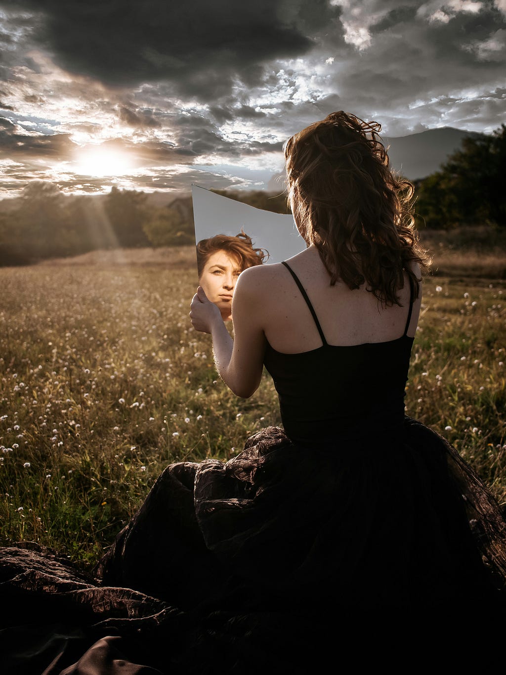 Woman sitting in field holding a mirror and looking at reflection