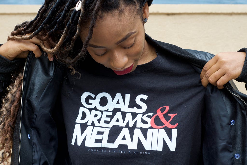 Young African-american woman showing a t-shirt that says: goals, dreams and melanin.