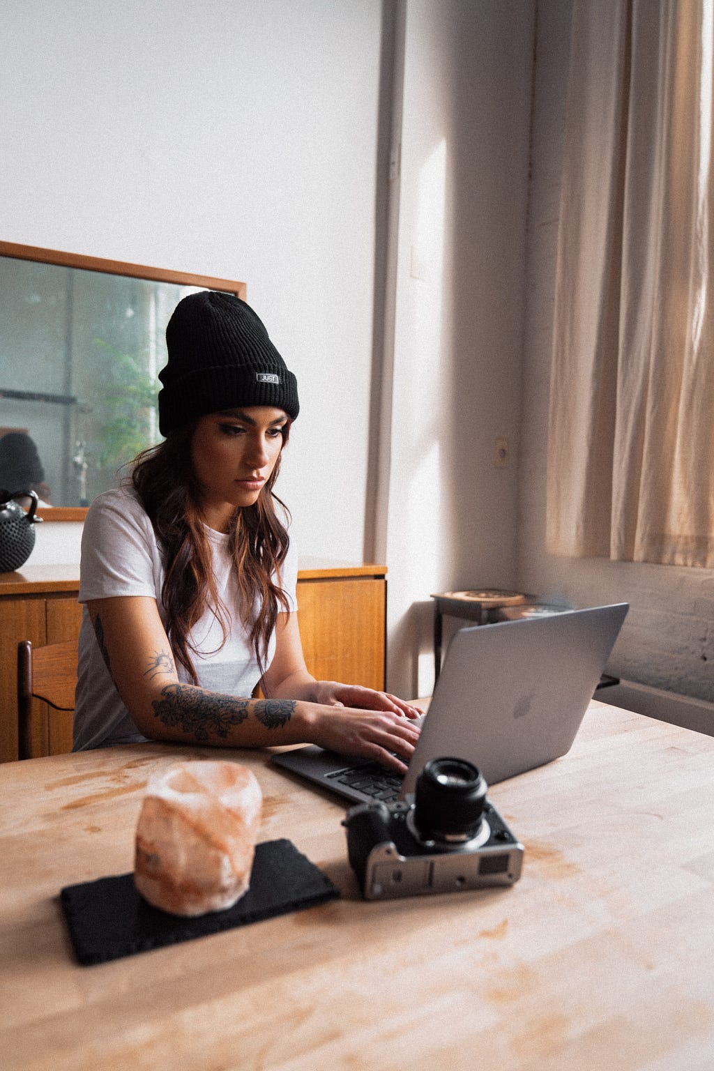 Young woman sitting at a table working on her laptop, with a camera laying on table nearby.