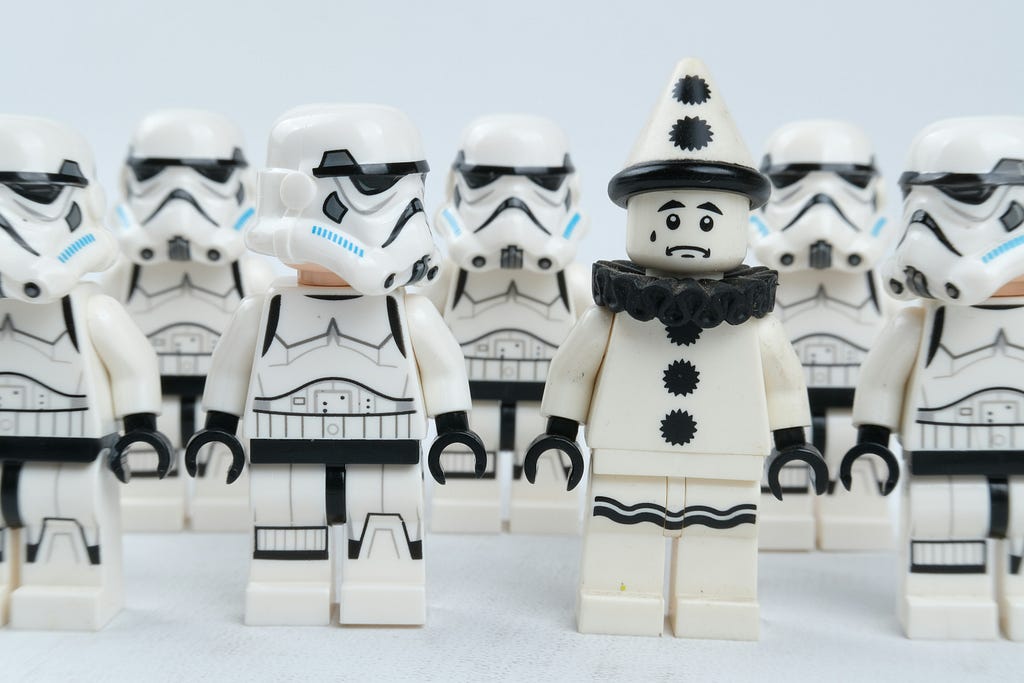 Foto: Eine Mannschaft schwarz-weiß uniformierter Lego-Klonkrieger aus der Star Wars Reihe steht in Reih und Glied. Dazwischen steht ein trauriger Clown in denselben Farbtönen. Die beiden nebenstehenden Klonkrieger haben jeweils ihre Köpfe in Richtung des CLowns gewandt.