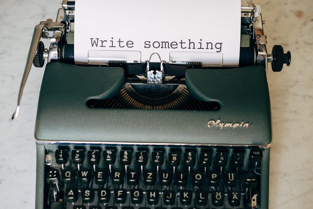 Uma máquina de escrever antiga sobre uma bancada de madeira. Há um papel na máquina datilografado com o seguinte dizeres em inglês: "Write something".