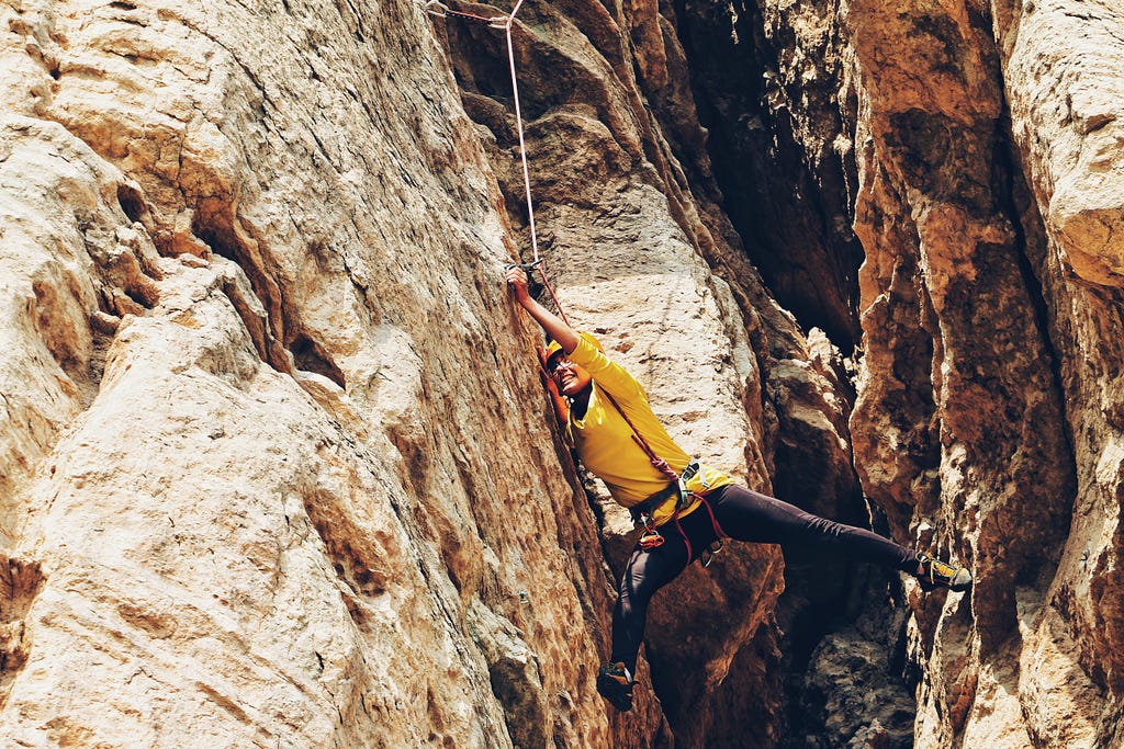 A woman climbing up a rocky cliff hangs from a rope, her face gritted with determination.