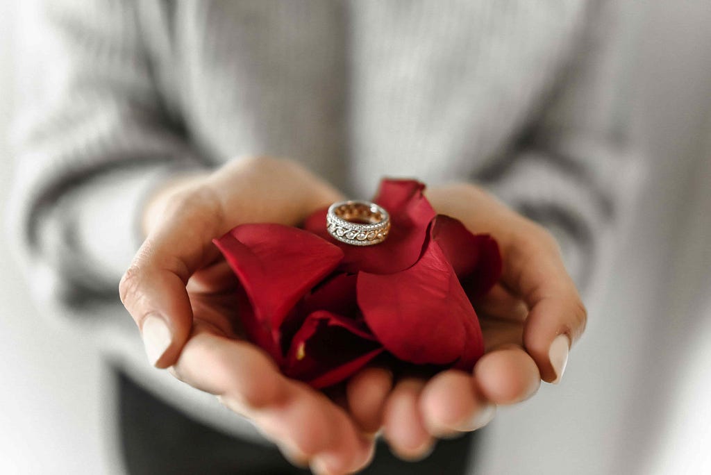 A beautiful engagement ring sitting on top of red rose petals, in the palms of someones hands.