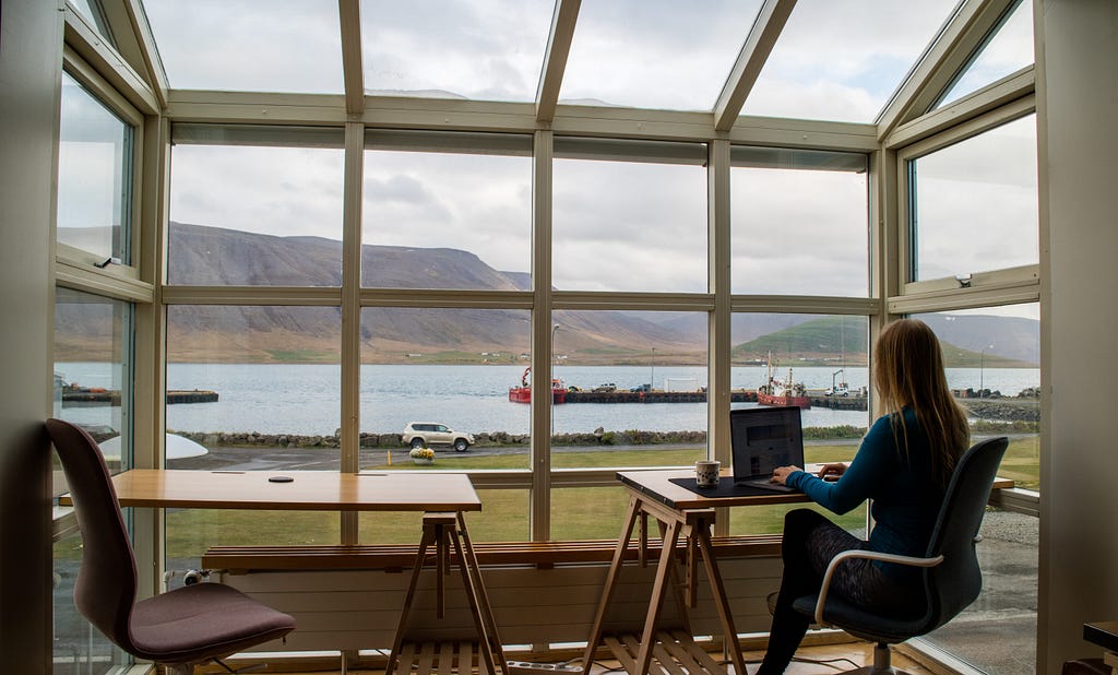 A woman siting at a sleek ergonomic desk typing on her laptop in front of a picture window overlooking a body of water and small mountain