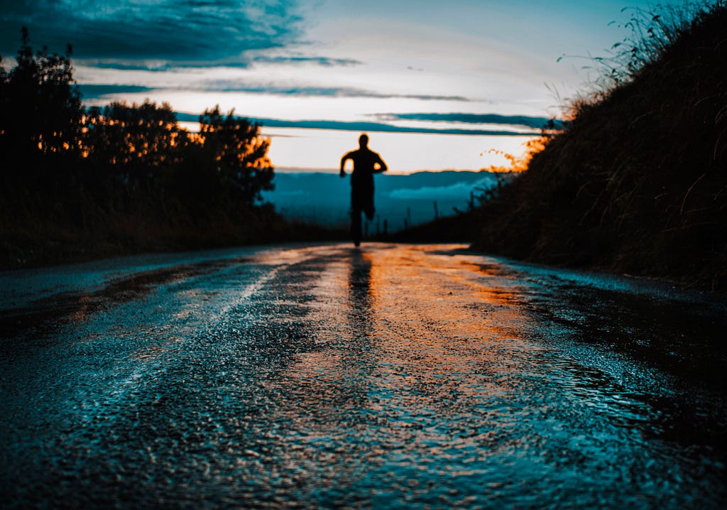 A shadowed figure is running down a road towards the sunset. Dark trees are on either side.