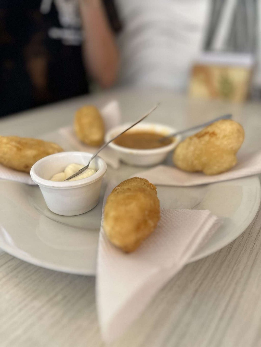 A plate with fried cassava fritters served on napkins, accompanied by small cups of dipping sauces with spoons, placed on a light-colored table.