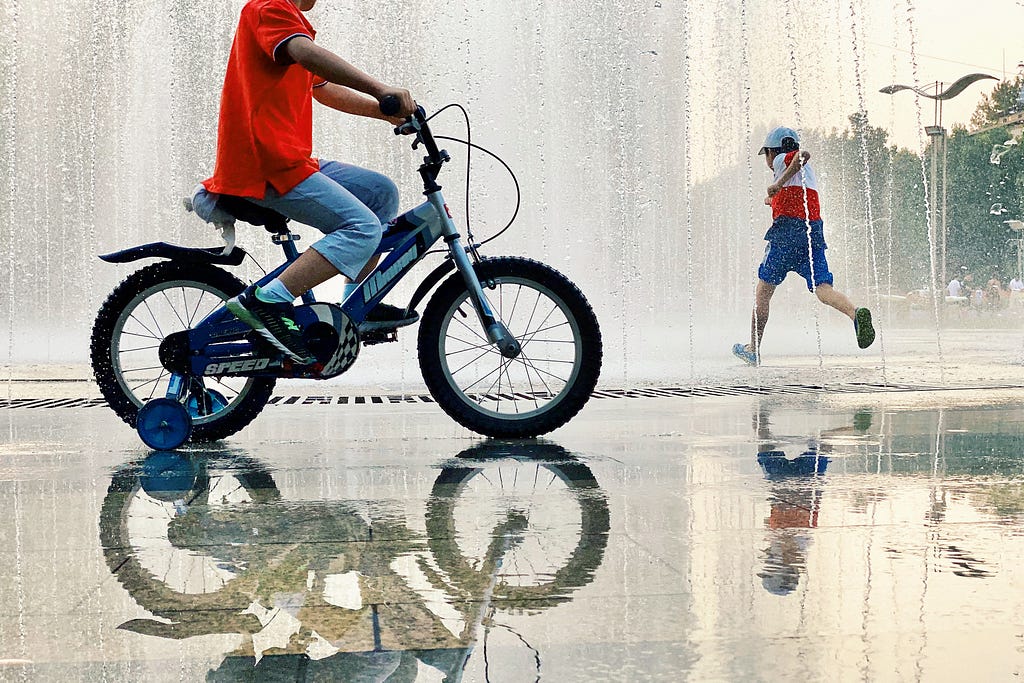 A child riding a bike with training wheels through sprinklers, and another child running in the distance.