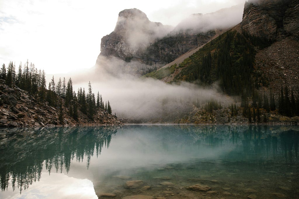 A lake surrounded by trees, all sitting in front of a mountain. The colors are dim and the sky is foggy, slightly obscuring the landscape.