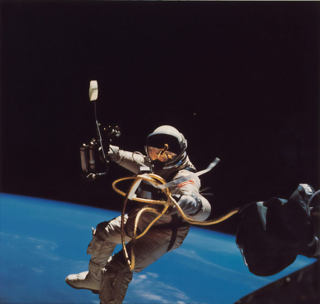 A person in a space suit holding a large tool is floating above the earth, tethered to a space craft with by a twisted cord.