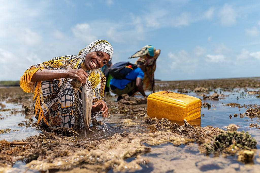 Amina stands waist deep in water off the coast of Lamu county, Kenya. She smiles, holding a small octopus in her hand. Two more octopus fishers crouch in the background.