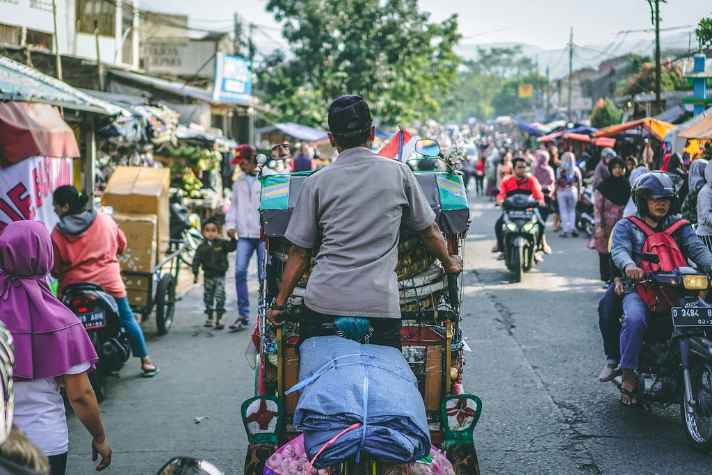 A photo of a rickshaw driver riding his rickshaw in the middle of a busy farmers market taken from behind.