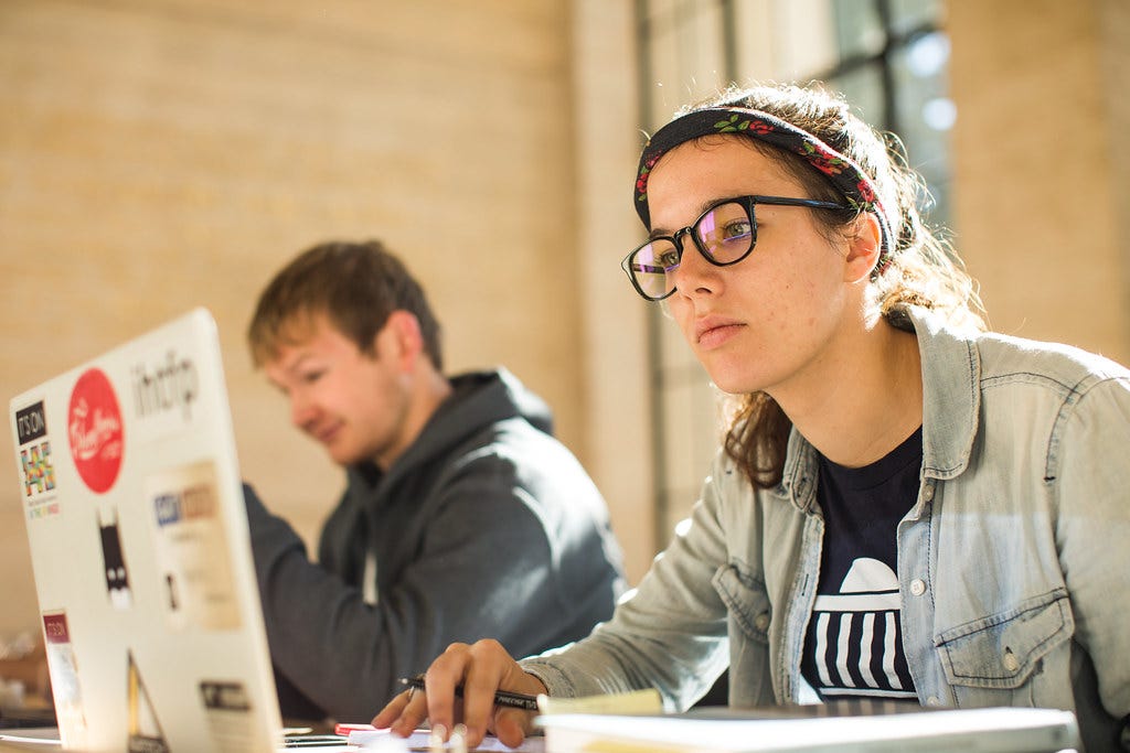 A young woman wearing glasses works at a laptop, with a young man in the background next to her