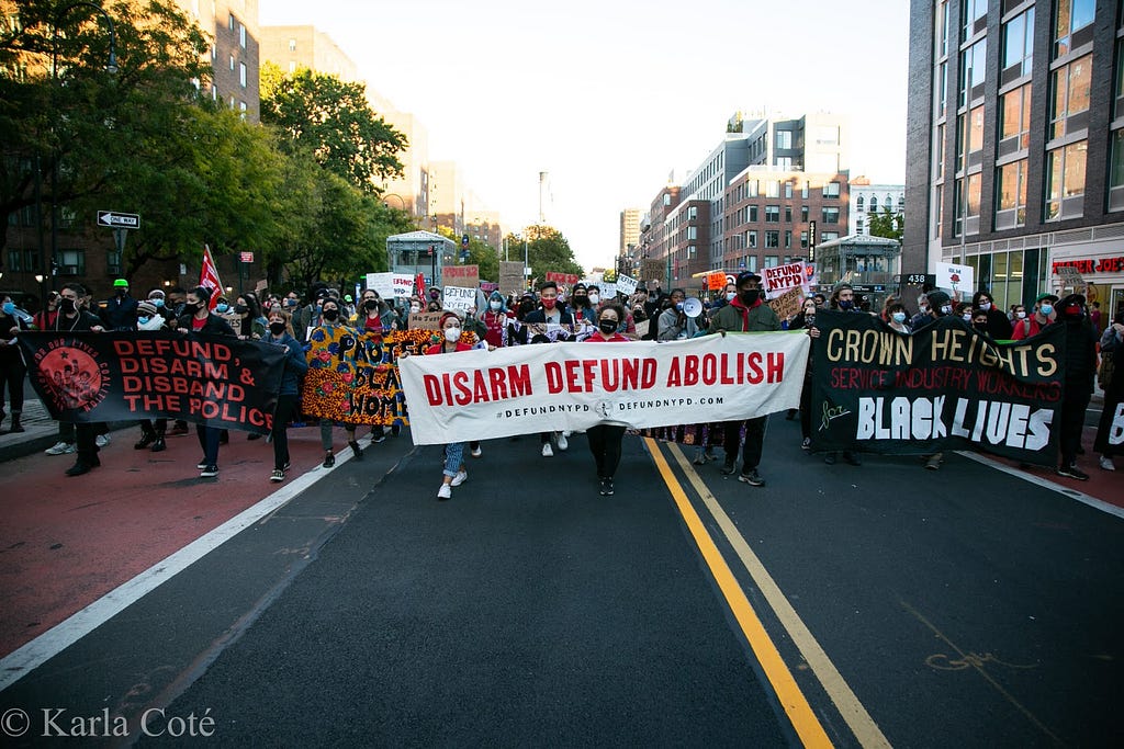 Image depicts hundreds of protesters gathered on October 17, 2020 to support the demand to defund the NYPD, tax the rich, and stop mass layoffs. The most prominent banners held by protesters in this image read “Defund, Disarm & Disband the Police,” “Protect Black Women,” “Disarm Defund Abolish | #DefundNYPD | DefundNYPD.com,” and “Crown Heights Service Industry Workers for Black Lives.”