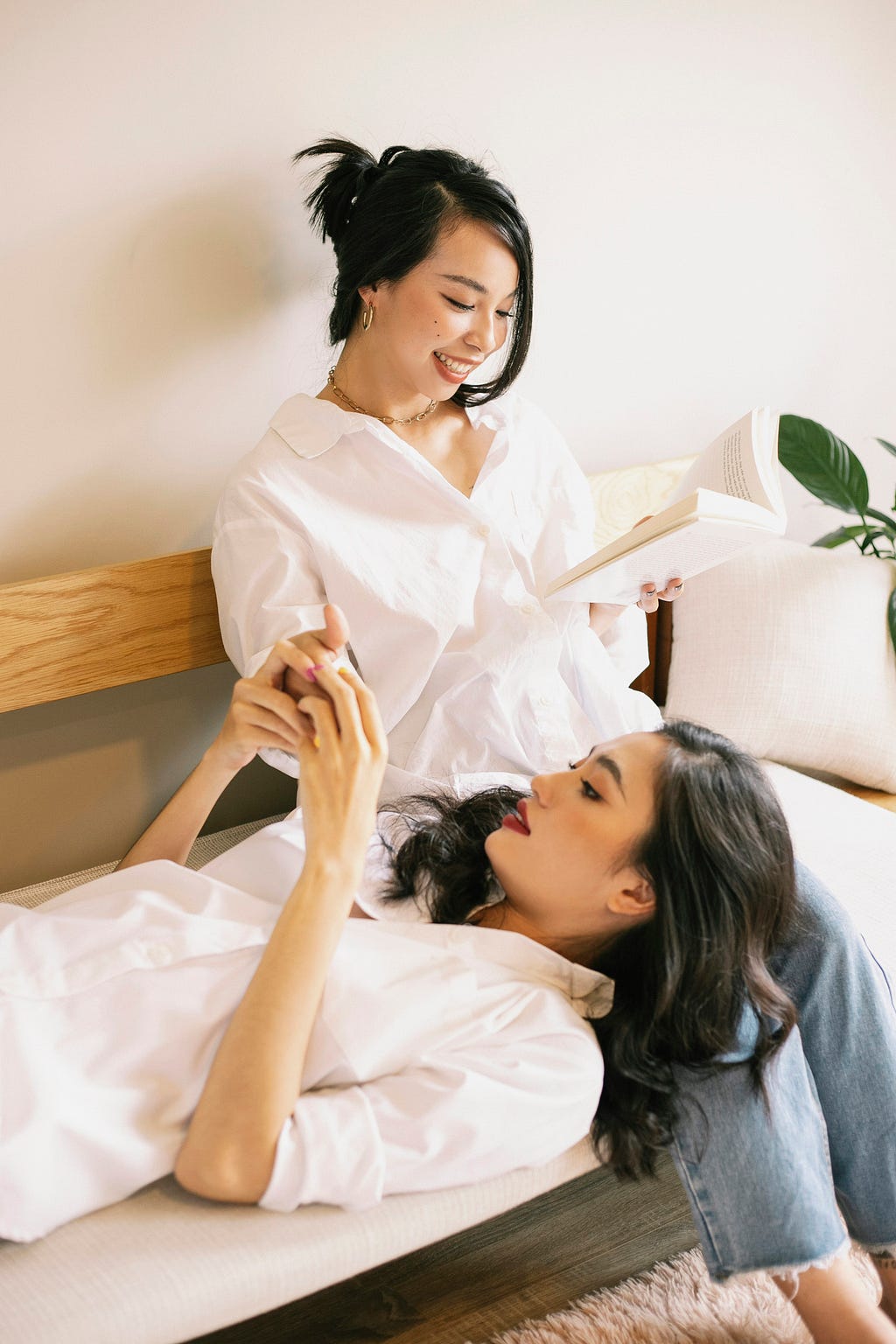 Two Asian women, one reading a book while the other lies with head in friend’s lap, holding her free hand