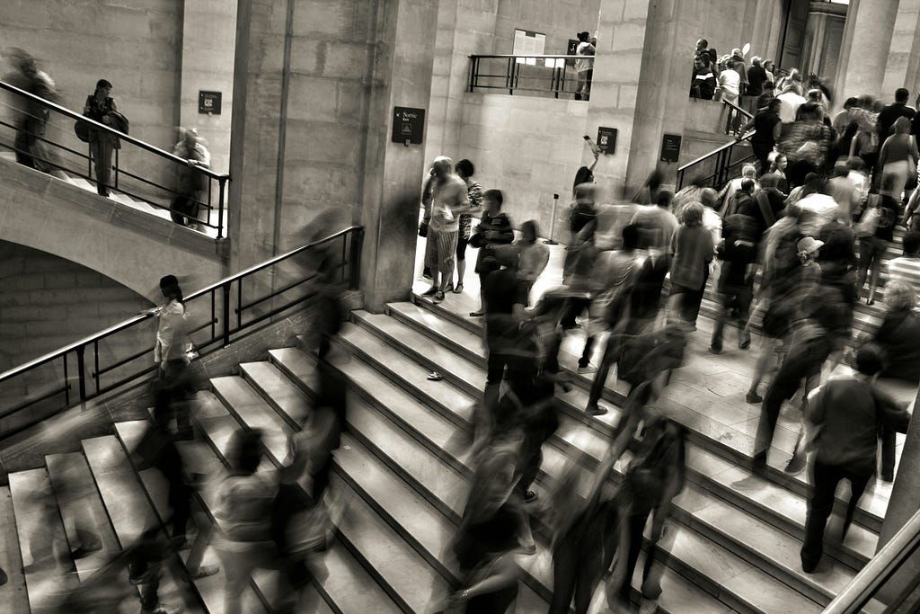 This image depicts a bustling interior scene, possibly within a museum or public building, characterized by a sense of motion and activity. The central focus is a grand staircase populated by numerous individuals depicted in various states of motion blur, which suggests the dynamic flow of people moving up and down. Some individuals are captured in stillness, offering contrast to the blurred motion around them, which gives the scene a temporal dimension. The architecture features classic stone w