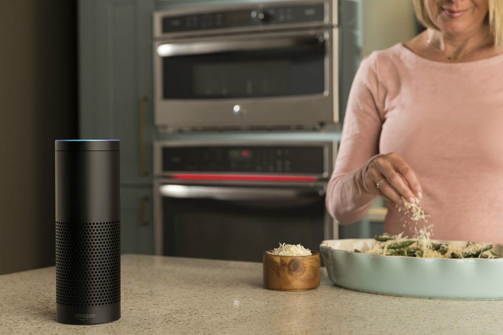An Alexa smart speaker sits on a kitchen worktop. In the background, a lady in a soft-pink cardigan makes dinner