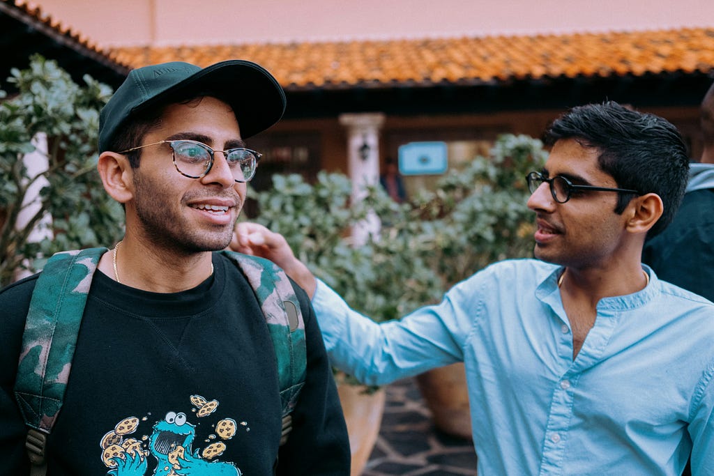 A color photo of two men from the chest up. They are casual looking and seem to be happy. One has his hand on the shoulder of the other in a friendly manner. The background is green, leafy bushes in front of the porch of a house with tile roofing.
