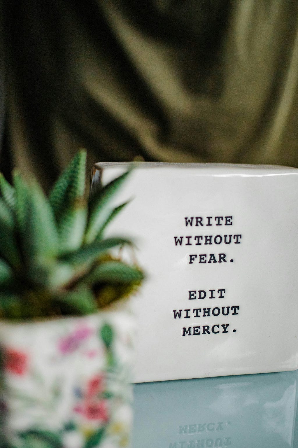 A color photograph — The backdrop is a olive green drape, a potted plant is in the foreground, the focus is a typewritten sign that states in all caps: WRITE WITHOUT FEAR. EDIT WITHOUT MERCY.