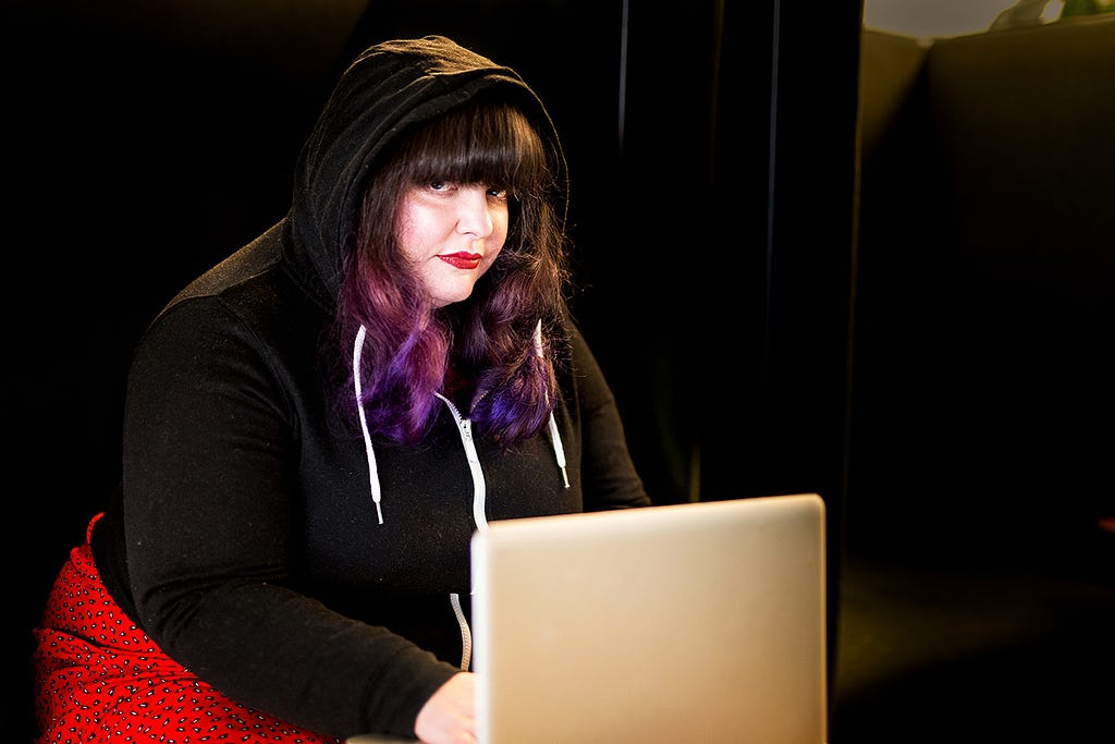 person with purple dyed hair and wearing red lipstick and a black hoodie sitting in front of a laptop