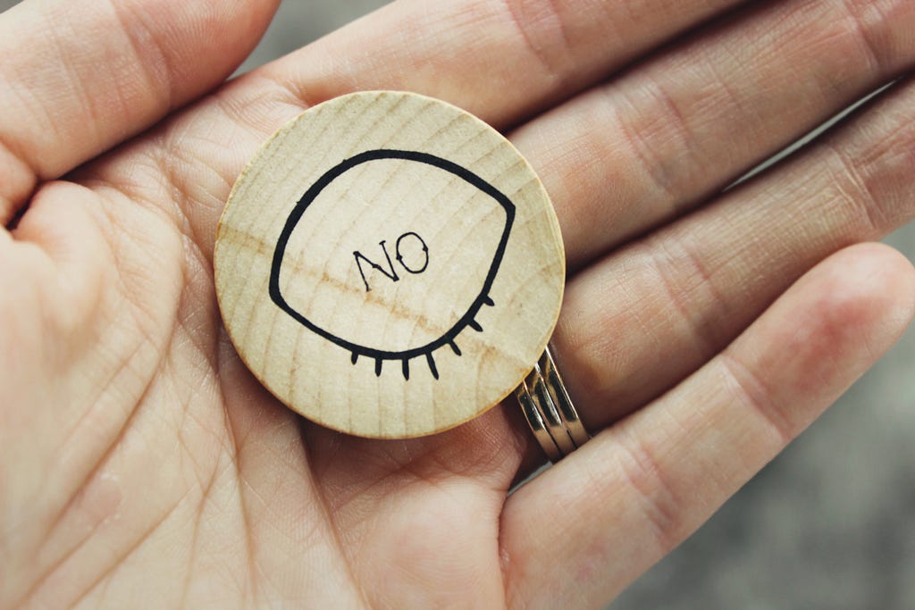 A hand holding a wooden coin that reads “No”