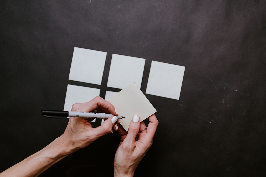 5 white post-its on a black wall, sixth post-it in a hand, a pen in another hand about to write on that post-it