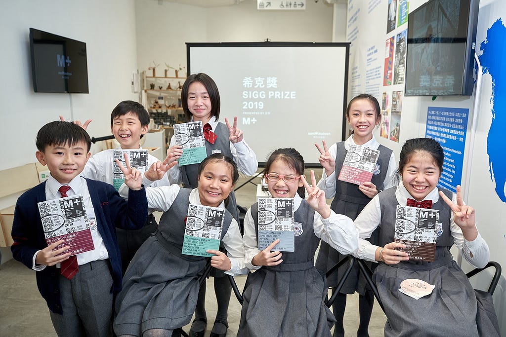 Seven smiling young students inside the M+ Pavilion pose and make peace signs. They each hold up an exhibition booklet.