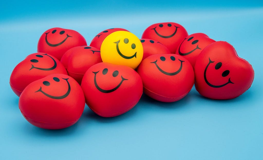 A yellow smiley ball sits among a group of red hearts against a light blue background, photo by Count Chris