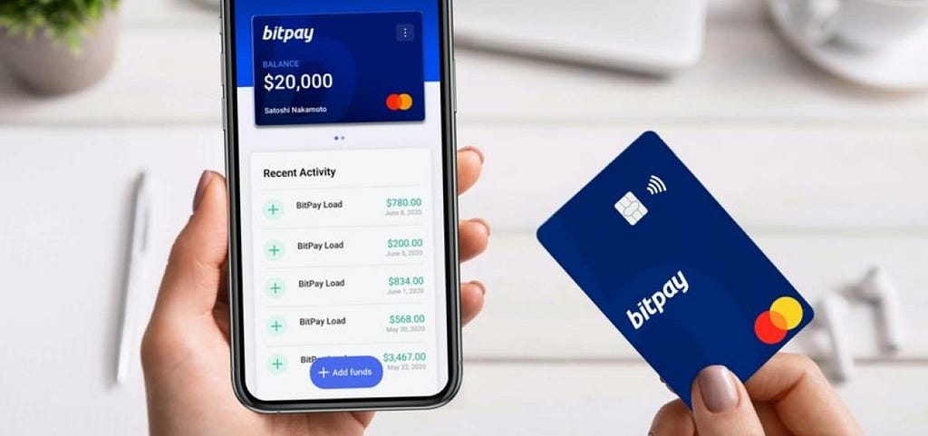 A woman holding a mobile phone with the Bitpay app open, displaying recent transactions. A debit card is held next to the device as a prop.