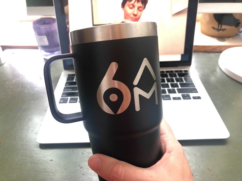 A hand holds a black Yeti travel mug with handle, branded with the 6AM City logo in silver lettering. Behind the mug, a laptop on video (showing one person) sits on a green desk.